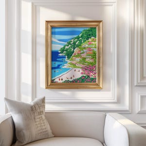 Beach art print with colorful flowers and bright houses and pink and green flowers on a mountainside hill with bright blue sky and little people on the shore and swimming in the bluey green water ocean sea. Calming and whimsical image of visitors to Positano village in Italy Europe. Thick heavy weight Gold Framed image of Positano hanging up against a White decorative wall in European architecture interior building or home with white couch and grey cushion below