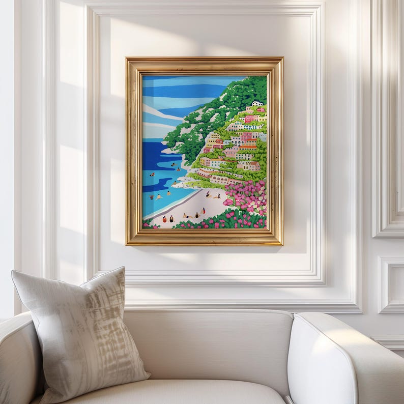 Beach art print with colorful flowers and bright houses and pink and green flowers on a mountainside hill with bright blue sky and little people on the shore and swimming in the bluey green water ocean sea. Calming and whimsical image of visitors to Positano village in Italy Europe. Thick heavy weight Gold Framed image of Positano hanging up against a White decorative wall in European architecture interior building or home with white couch and grey cushion below