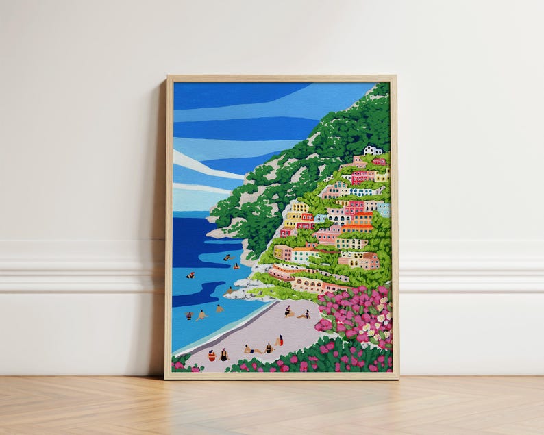 Beach art print with colorful flowers and bright houses and pink and green flowers on a mountainside hill with bright blue sky and little people on the shore and swimming in the bluey green water ocean sea. Calming and whimsical image of visitors to Positano village in Italy Europe. Neutral wooden soft light framed image of Positano up against a White decorative wall in European architecture interior building sitting on soft wooden herringbone floors