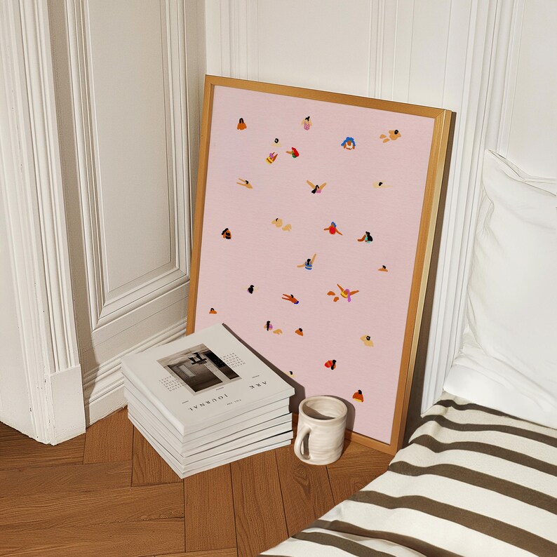 May include: A framed print of a pink background with small, colorful illustrations of people in various swimming positions. The print is leaning against a stack of white journals with the text "2022 JOURNAL" printed on the top journal.