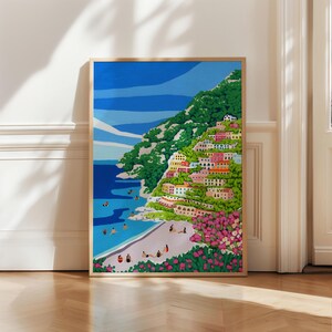 Beach art print with colorful flowers and bright houses and pink and green flowers on a mountainside hill with bright blue sky and little people on the shore and swimming in the bluey green water ocean sea. Calming and whimsical image of visitors to Positano village in Italy Europe. Neutral wooden soft light framed image of Positano up against a White decorative wall in European architecture interior building sitting on soft wooden herringbone floors