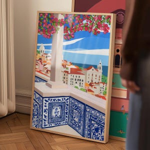 May include: A framed print of a colorful illustration depicting a cityscape with a blue tiled balcony overlooking the ocean. The cityscape features buildings with red roofs and a tall white tower. Pink flowers are blooming on a tree in the foreground.