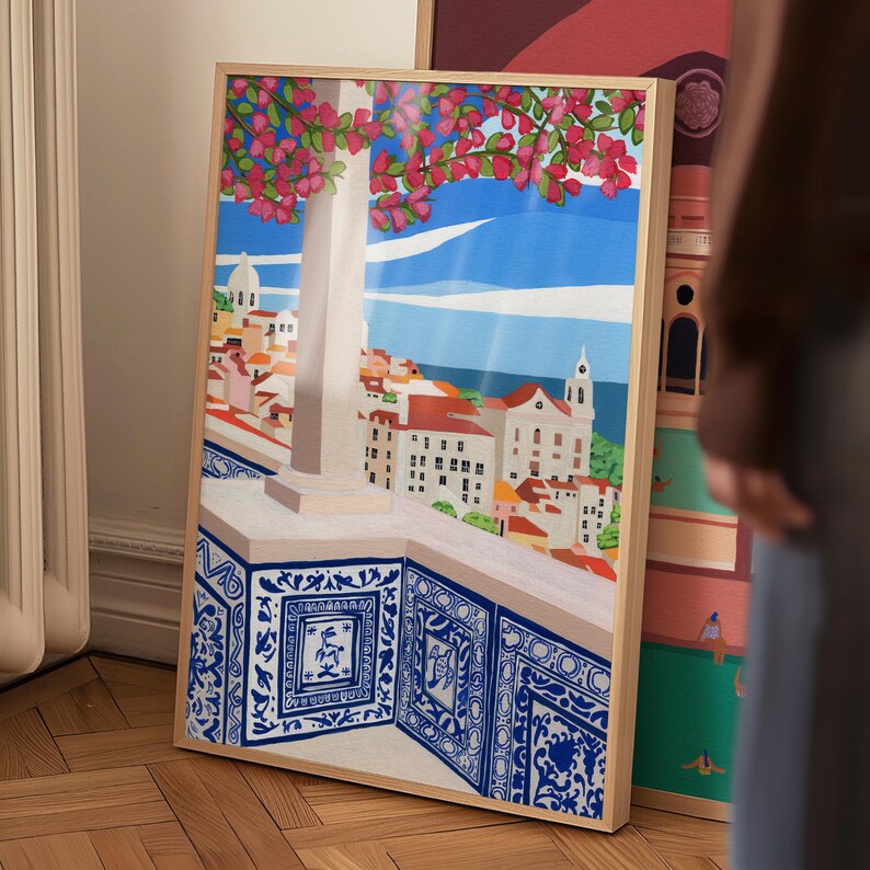 May include: A framed print of a colorful illustration depicting a cityscape with a blue tiled balcony overlooking the ocean. The cityscape features buildings with red roofs and a tall white tower. Pink flowers are blooming on a tree in the foreground.