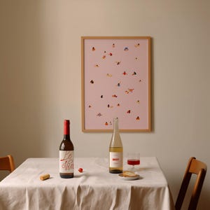 May include: A white tablecloth covers a table with two bottles of wine, one red and one white, a glass of red wine, and a piece of bread. A framed print of a pink background with small figures of people in various poses hangs on the wall behind the table.
