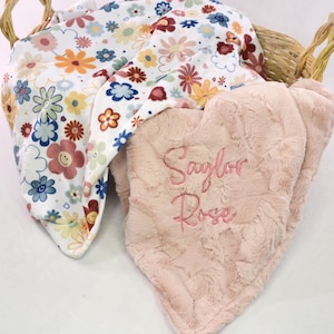 May include: A woven basket holds two baby blankets. One blanket has a floral pattern with yellow, orange, blue, and brown flowers on a white background. The other is a soft, pink blanket with the embroidered name "Saylor Rose".