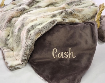 Puede incluir: Una manta de piel sintética en una cesta. La manta presenta una mezcla de tonos blancos, marrones y beige. Una almohada de terciopelo marrón oscuro está bordada con el nombre "Cash" en cursiva. La cesta tiene un asa tejida.