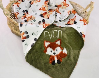May include: A baby blanket with a fox theme. The blanket features a white background with orange foxes and green leaves. The blanket is folded over a green blanket with a fox applique and the name "Evan". The blanket is in a wicker basket.