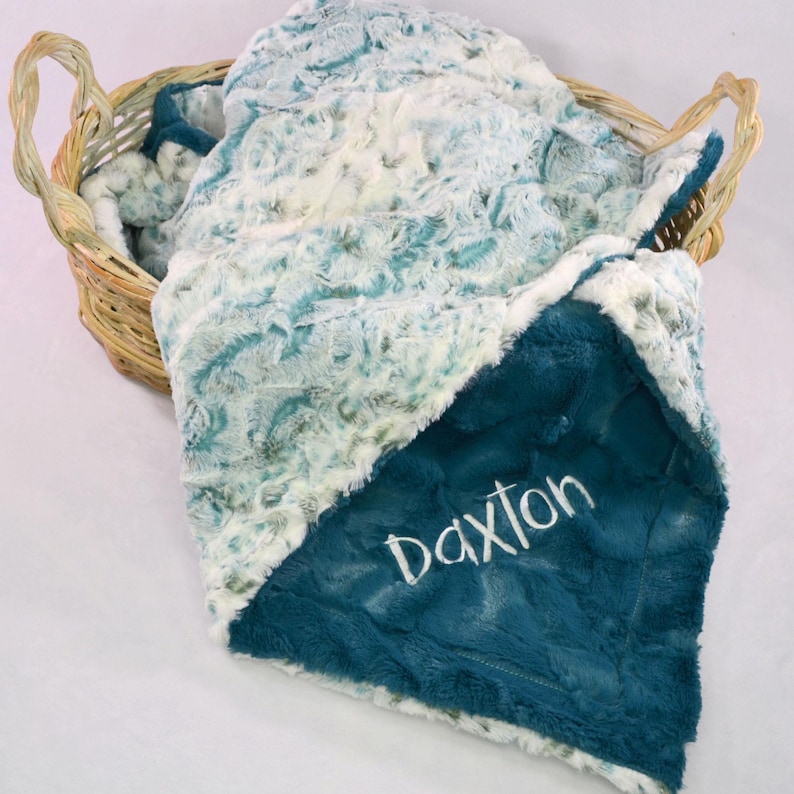 May include: A soft, plush baby blanket with a teal blue side and a white side with a teal blue swirl pattern. The teal blue side has the name "Daxlon" embroidered in white.