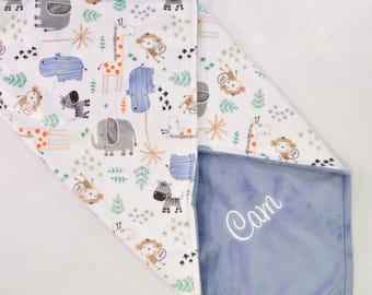 May include: A white and blue baby blanket with a jungle animal print. The blanket is folded in half and the side with the animal print is visible. The other side of the blanket is a solid blue colour with the name "Cam" embroidered in white.