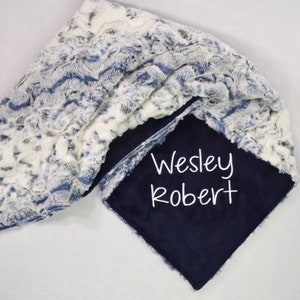 May include: A soft, plush blanket with a navy blue backing and a white and blue patterned top. The blanket is personalized with the name "Wesley Robert" in white script. The blanket is made of a soft, textured material.