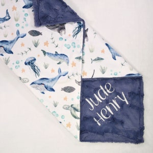 May include: A white and blue baby blanket with a sea life pattern featuring whales, dolphins, jellyfish, and starfish. The blanket has a soft, blue minky backing with the names "Jude Henry" embroidered in white.