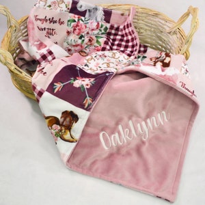May include: A pink and white patchwork baby blanket with a floral and horse design. The blanket is personalized with the name "Oaklynn" embroidered in white on the pink side.