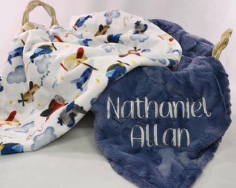 May include: A blue and white baby blanket with a pattern of aeroplanes and clouds. The blanket is personalised with the name "Nathaniel Allan" in white embroidery.