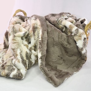 May include: A soft, plush throw blanket with a faux fur texture. One side is a light brown with a white pattern, and the other side is a solid light brown.