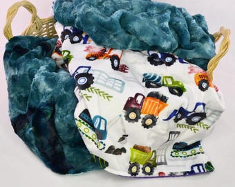May include: A soft, plush blanket with a teal blue side and a white side with a colorful construction vehicle print. The blanket is folded and placed in a wicker basket.