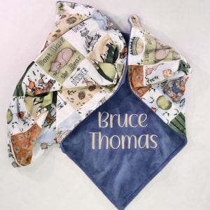 May include: A blue and patchwork baby blanket with the embroidered name "Bruce Thomas." The patchwork features woodland animal illustrations and the phrases "Don't Wake the Bear," "Dream," and "Discover."