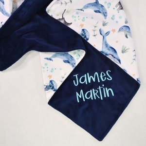 May include: A white and blue baby blanket with a navy blue fleece backing. The blanket features a whimsical ocean-themed print with whales, dolphins, jellyfish, and seaweed. The name "James Martin" is embroidered in turquoise on the navy blue side.