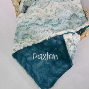 May include: A teal blue and white baby blanket with a soft, plush texture. The blanket is personalized with the name "Daxon" embroidered in white.