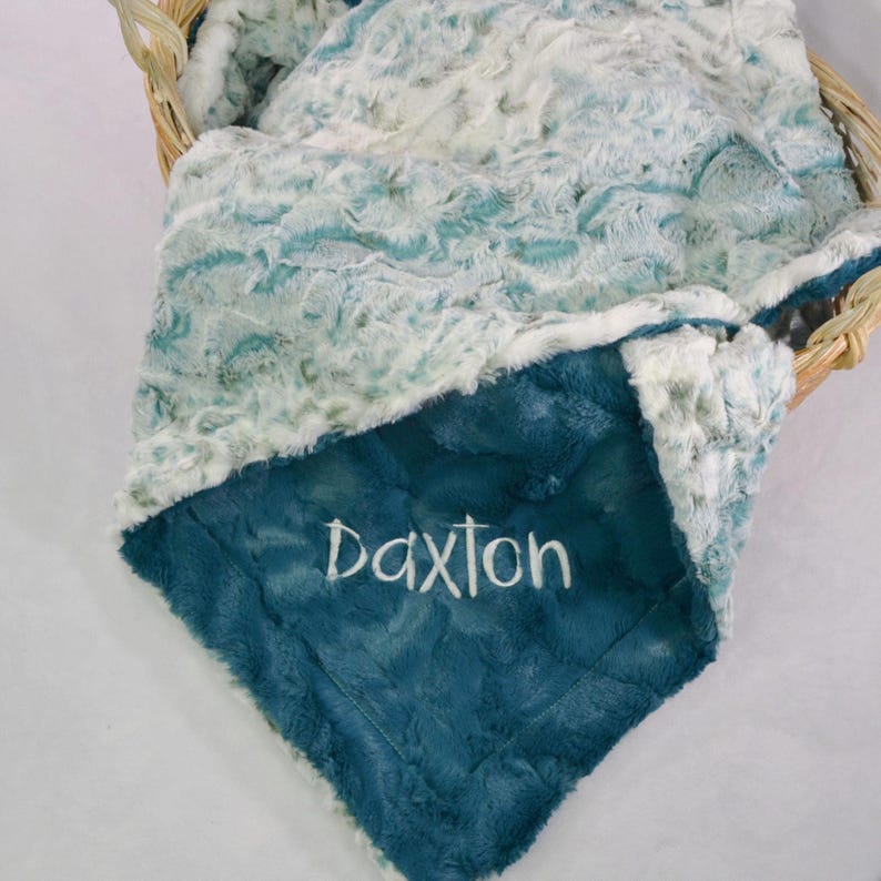 May include: A teal blue and white baby blanket with a soft, plush texture. The blanket is personalized with the name "Daxon" embroidered in white.