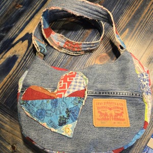 May include: A denim shoulder bag with a patchwork heart applique. The bag features a Levi Strauss & Co. label and a patterned strap. The bag is made from recycled denim and fabric scraps.