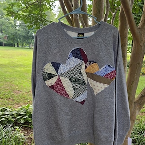 May include: A heather gray long-sleeve sweatshirt featuring a large, central appliqué of two overlapping hearts. The hearts are constructed from various fabric scraps in diverse patterns and colors, including floral, plaid, and geometric prints.