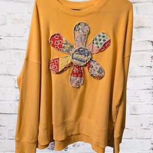 May include: A mustard yellow long-sleeve sweatshirt featuring a large, central flower appliqué. The flower is constructed from various fabric patches with diverse patterns, including red geometric shapes, blue polka dots, and plaid textures.