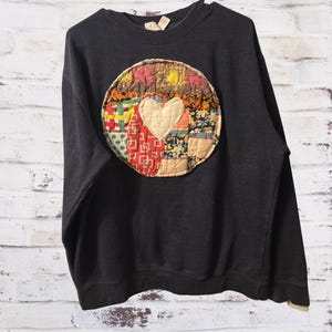 May include: A charcoal gray crewneck sweatshirt featuring a large, circular, quilted patch on the chest. The patch is composed of various colorful fabric squares in red, yellow, and blue, with a central cream-colored heart shape stitched on top.