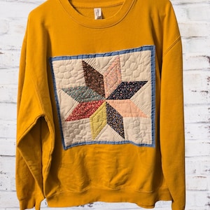 Könnte beinhalten: Ein senfgelbes Langarm-Sweatshirt mit einem großen, quadratischen, gesteppten Aufnäher auf der Vorderseite. Der Aufnäher zeigt ein mehrfarbiges Sternmuster aus verschiedenen floralen und einfarbigen Stoffstücken in Braun-, Rosa-, Blau- und Gelbtönen.