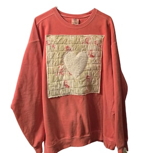 May include: A coral-colored long-sleeve crewneck sweatshirt featuring a large, square, quilted patch on the front. The patch has a light yellow background with a pink floral pattern and a central white heart shape with visible stitching.