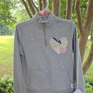 May include: A heather gray long-sleeve pullover with a quarter-zip neckline and two front pockets. The chest features a large, quilted heart appliqué made from various patterned fabric scraps, with a smaller solid pink heart attached to it.