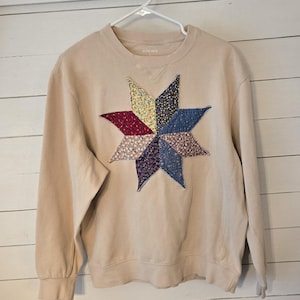 May include: A beige crewneck sweatshirt featuring a large, eight-pointed star appliqué on the chest. The star is constructed from various floral-patterned fabric patches in shades of yellow, red, blue, and purple, with visible stitching.