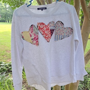 May include: A light gray heathered long-sleeve sweatshirt featuring three heart-shaped fabric appliqués across the chest. Each heart is crafted from different floral and patterned fabric scraps with raw, dark edges. The inner label reads ocean drive.