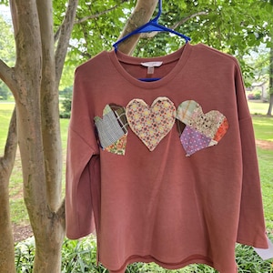 May include: A dusty rose long-sleeve sweatshirt featuring three heart-shaped fabric appliqués across the chest. Each heart is constructed from a patchwork of various patterned fabrics, including plaid, floral, and geometric prints in multiple colors.