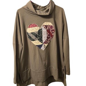 May include: A tan long-sleeve pullover sweater with a cowl neckline and a high-low hem. The center of the chest features a large heart-shaped appliqué constructed from various pieces of fabric in floral, plaid, and solid patterns in shades of red, blue, and cream.