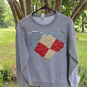 Puede incluir: Una sudadera gris jaspeado de manga larga y cuello redondo con una gran aplicación en forma de corazón en el centro. El corazón está confeccionado con parches de tela acolchada en tonos azul claro, crema con flores y rojo.