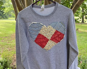 May include: A heather gray long-sleeve crewneck sweatshirt featuring a large, central heart-shaped appliqué. The heart is constructed from quilted fabric patches in shades of light blue, cream with a small floral print, and solid red.
