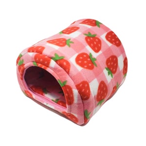 May include: A pink and white checkered pet bed with a strawberry pattern. The bed is made of soft fleece material and has a rounded shape with an opening for entry. The strawberries are red with green stems.