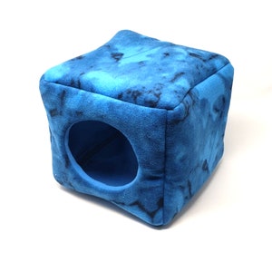 Puede incluir: Una cama para mascotas azul en forma de cubo con una entrada redonda. La cama tiene un patrón de teñido anudado en tonos azules y negros. El material suave y afelpado está diseñado para mascotas pequeñas.