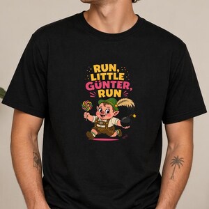 Könnte beinhalten: Schwarzes T-Shirt mit einem Cartoon-Kobold, der einen Lolli hält, mit dem Text "RUN, LITTLE GUNTER, RUN" in Pink und Gelb. Der Kobold trägt einen grünen Hut und ein braunes Outfit. Das T-Shirt ist aus weichem Material.