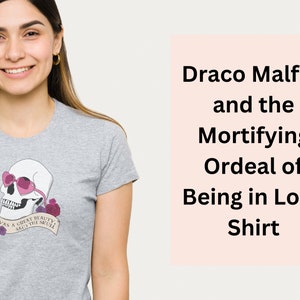 Könnte beinhalten: Graues T-Shirt mit einem rosafarbenen Schädel, der eine herzförmige Sonnenbrille trägt und ein Banner mit der Aufschrift "Twas a great beauty, says the skull". Der Text "Draco Malfoy and the Mortifying Ordeal of Being in Love Shirt" ist in schwarzer Schrift auf rosa Hintergrund.