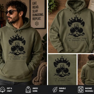 Könnte beinhalten: Olivgrüner Kapuzenpullover mit einem schwarzen Aufdruck einer Katze mit Krone und Sonnenbrille, mit dem Text "I've always been a badass, Carl." Der Hoodie hat eine Vordertasche und eine Kapuze mit Kordelzug.