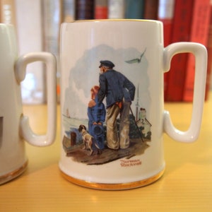 May include: Two white ceramic mugs with illustrations. The mug on the left features a sailor holding a rope. The mug on the right features a sailor with a child and a dog. The mug on the right has the text "Norman Rockwell" at the bottom.