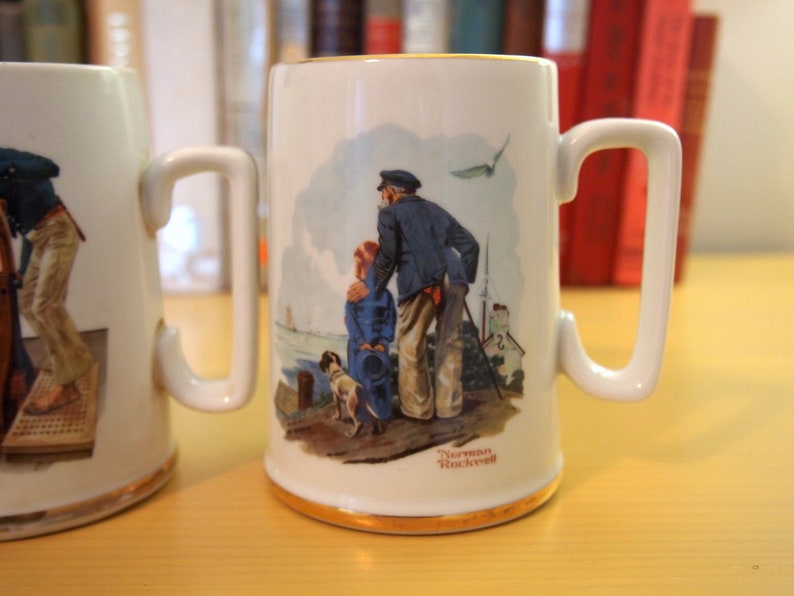 May include: Two white ceramic mugs with illustrations. The mug on the left features a sailor holding a rope. The mug on the right features a sailor with a child and a dog. The mug on the right has the text "Norman Rockwell" at the bottom.