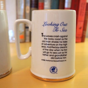 May include: White ceramic mug with a blue and gold trim. The mug has a handle and features a poem about looking out to sea. The poem reads "The waves crash against the rocky coast as the old man shares his tales of adventure on the high seas. And the boy dreams of the day when he too will go to sea just as his father and grandfather did before him." The mug also has a blue and white logo with the text "The Norman Rockwell Museum, Inc."