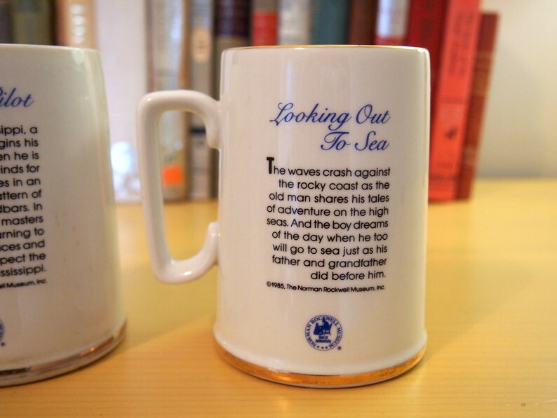May include: White ceramic mug with a blue and gold trim. The mug has a handle and features a poem about looking out to sea. The poem reads "The waves crash against the rocky coast as the old man shares his tales of adventure on the high seas. And the boy dreams of the day when he too will go to sea just as his father and grandfather did before him." The mug also has a blue and white logo with the text "The Norman Rockwell Museum, Inc."