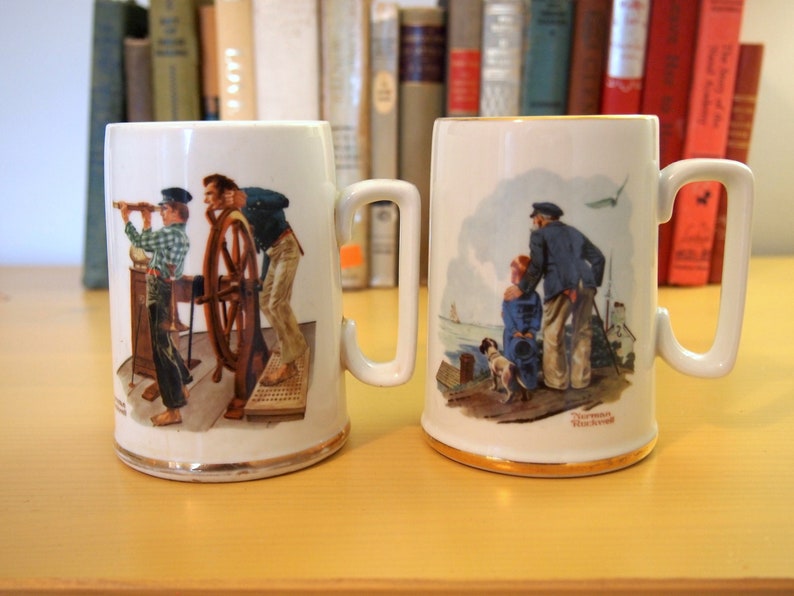 May include: Two white ceramic mugs with illustrations of Norman Rockwell paintings. The mug on the left features a man at the helm of a ship, while the mug on the right depicts a man and a child standing on a dock. Both mugs have gold trim around the rim.