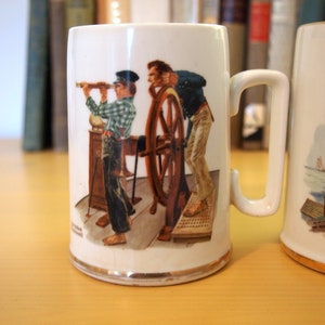 May include: Two white ceramic mugs with illustrations. The mug on the left features a scene of two men on a ship, one holding a telescope and the other steering a wheel. The mug on the right features a scene of a dog and a woman on a boat.