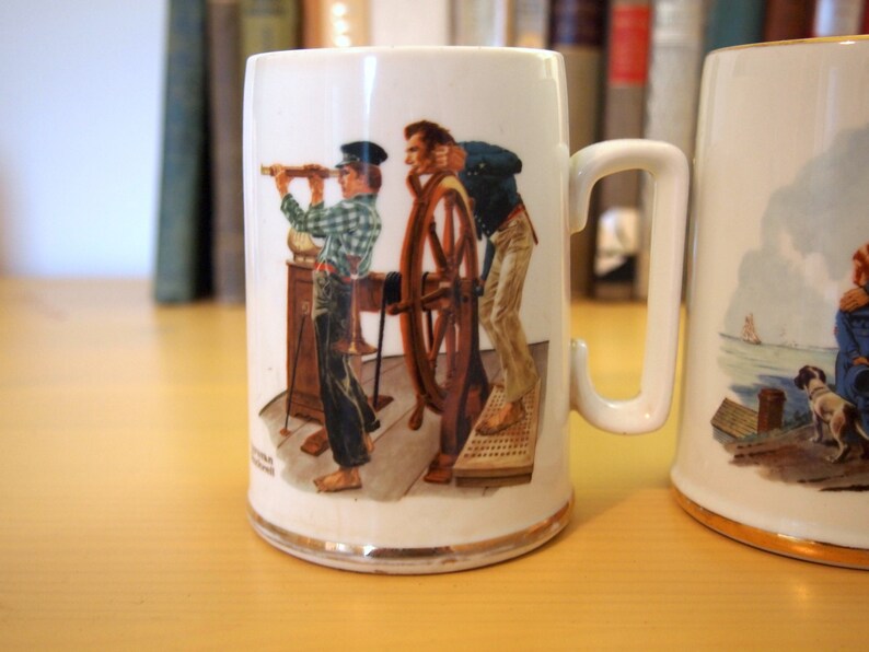 May include: Two white ceramic mugs with illustrations. The mug on the left features a scene of two men on a ship, one holding a telescope and the other steering a wheel. The mug on the right features a scene of a dog and a woman on a boat.