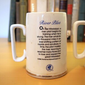 May include: White ceramic mug with a blue and gold design. The mug features a quote about river pilots on the Mississippi River. The quote reads: "On the Mississippi, a river pilot begins his training when he is young. The river winds for a thousand miles in an ever-shifting pattern of rapids and sandbars. In time, the pilot masters the river, learning to read her many faces and to love and respect the great Mississippi." The mug is also decorated with a gold rim and a small blue and gold emblem.