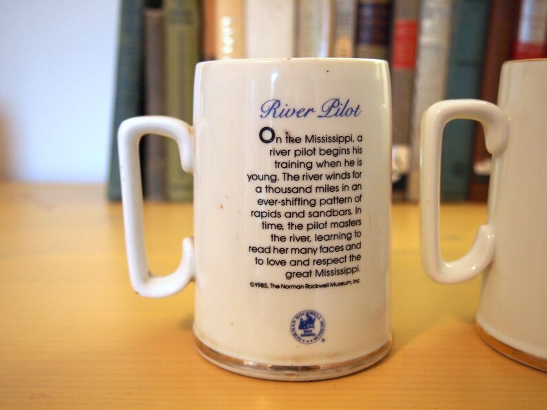 May include: White ceramic mug with a blue and gold design. The mug features a quote about river pilots on the Mississippi River. The quote reads: "On the Mississippi, a river pilot begins his training when he is young. The river winds for a thousand miles in an ever-shifting pattern of rapids and sandbars. In time, the pilot masters the river, learning to read her many faces and to love and respect the great Mississippi." The mug is also decorated with a gold rim and a small blue and gold emblem.